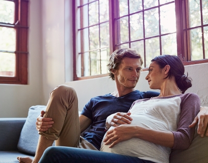 Mann und Frau sitzen gemeinsam auf dem Sofa. Sie ist schwanger. #Gib8aufGrippeschutz