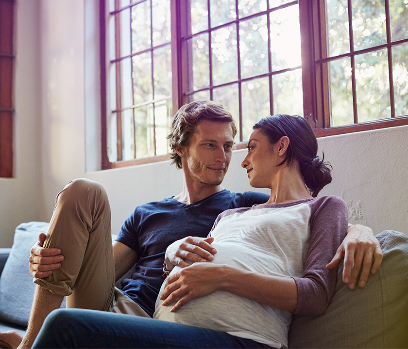 Mann und Frau sitzen gemeinsam auf dem Sofa. Sie ist schwanger. #Gib8aufGrippeschutz