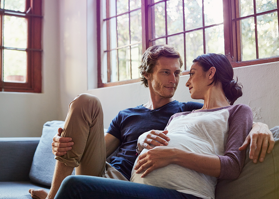 Mann und Frau sitzen gemeinsam auf dem Sofa. Sie ist schwanger. #Gib8aufGrippeschutz.